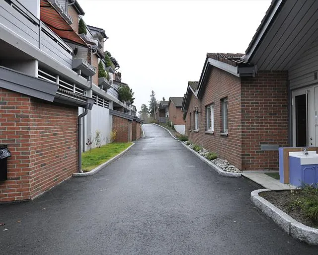 Brolagt gangvei mellom rekkehus og leiligheter med murfasader og grønne planter.