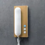Dørtelefon med front i lyst treverk, hvitt telefonrør og tre knapper, montert på grå vegg.