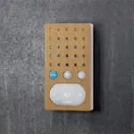 Intercom i lyst treverk-design med hvit høyttalerdel og tre knapper, på mørk bakgrunn.