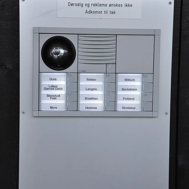 Dørtelefon med kamera, høyttaler og navneskilt montert på mørk vegg, med skilt mot reklame.