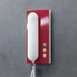 Rød dørtelefon med hvitt rør og tre knapper, på mørk vegg.