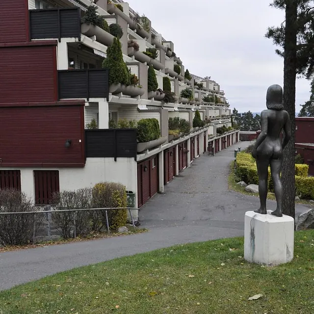 Terrasseblokker med beplantede balkonger i flere etasjer, sett fra gangvei med statue i forgrunnen.