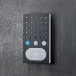 Mørk intercom-enhet med teksturert overflate, hvit høyttalerdel og tre knapper, på mørk vegg.