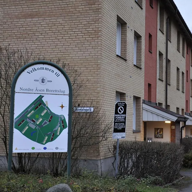 Skilt med kart og tekst "Velkommen til Nordre Åsen Borettslag" foran mursteinsbygning.
