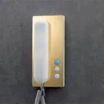 Gullfarget dørtelefon med hvitt rør og tre knapper, montert på grå vegg.