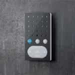 Mørk grå intercom-enhet med hvit høyttalerdel og tre knapper, montert på mørk vegg.