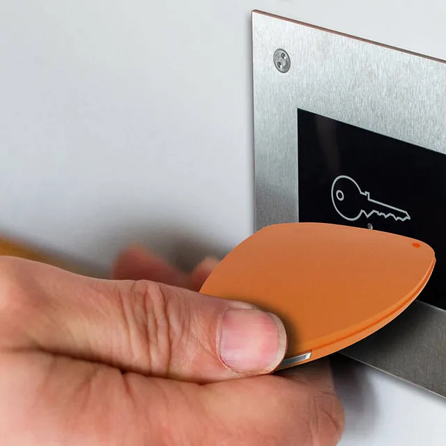 En hånd holder en oransje nøkkelbrikke foran en elektronisk kortleser med nøkkelsymbol.