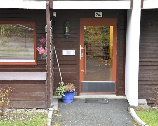 Inngangsdør merket 24B i brun trebygning, med ringetablå og blomsterpotte ved siden.