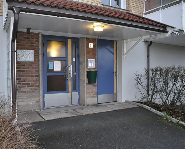 Inngangsparti med blå dør og ringetablå i boligblokk med mursteinsfasade og overbygg.