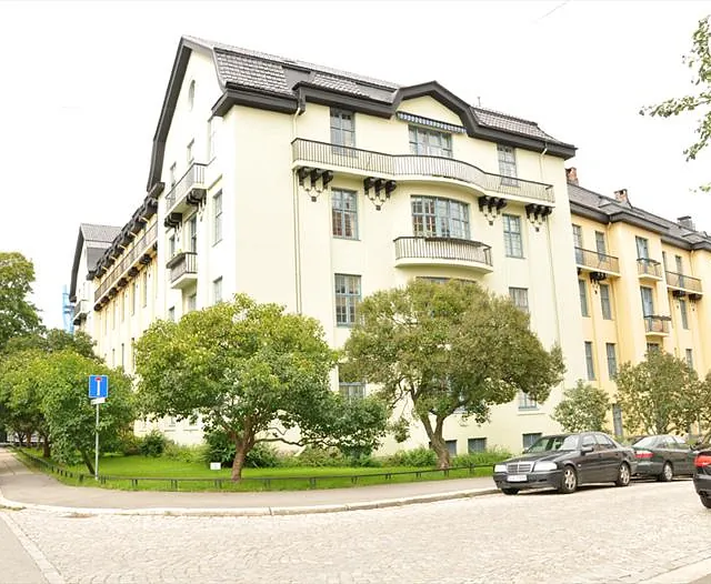 Stor, lys gul bygård med mørkt tak, balkonger og trær foran på grøntområde.