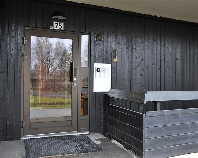 Inngangsparti med mørk trepanel, glassdør merket med nummer 75 og dørtelefon til høyre.