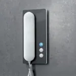 Grå dørtelefon med hvit telefonrør og tre knapper, montert på grå vegg.