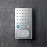 Sølvfarget intercom med perforert front, tre knapper og hvit høyttalerdel, på mørk vegg.