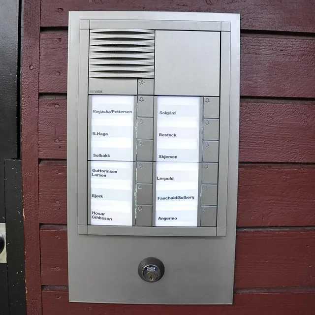 Nærbilde av ringetablå med navn og ringeknapper, montert på rød panelvegg ved inngangsdør.