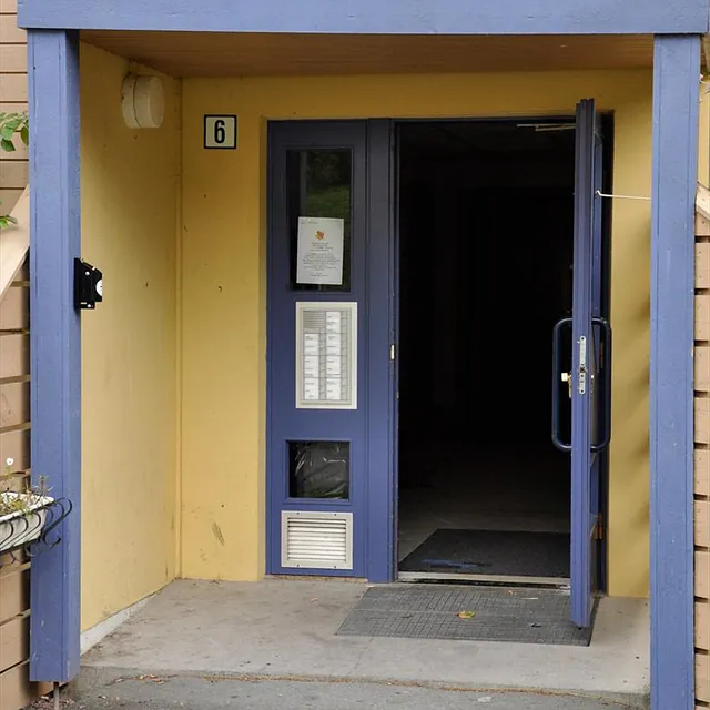 Inngangsparti med blå dør i gul vegg, dørtelefon og navneliste montert ved siden av inngangen.